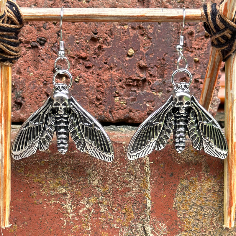 Death's-head moth dangle earrings in silver-tone metal on fish-hook wires, displayed on a wooden stand against a brick background